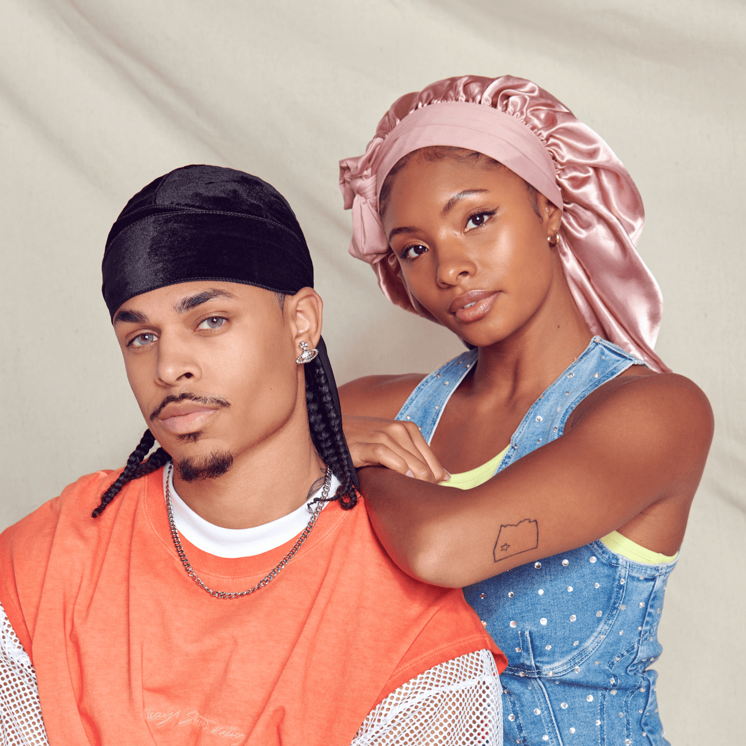 Two people posing together against a neutral background; man in orange shirt with black headband, woman in denim vest and...