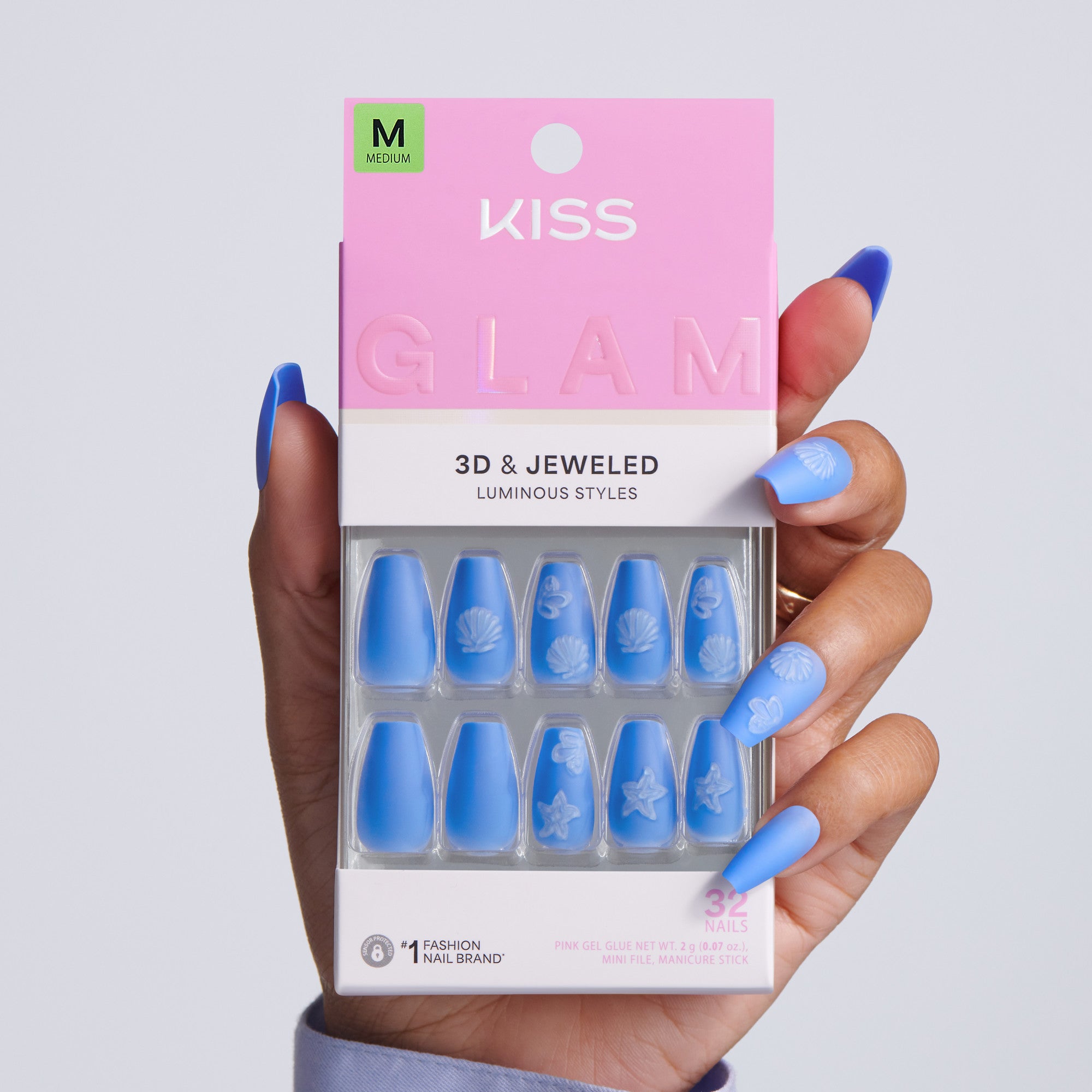 Hand holding KISS Glam 3D &amp; Jeweled Luminous Styles press-on nails packaging, Medium size, 32 nails, blue medium coffin nails with white embossed shell and ocean designs on gray background