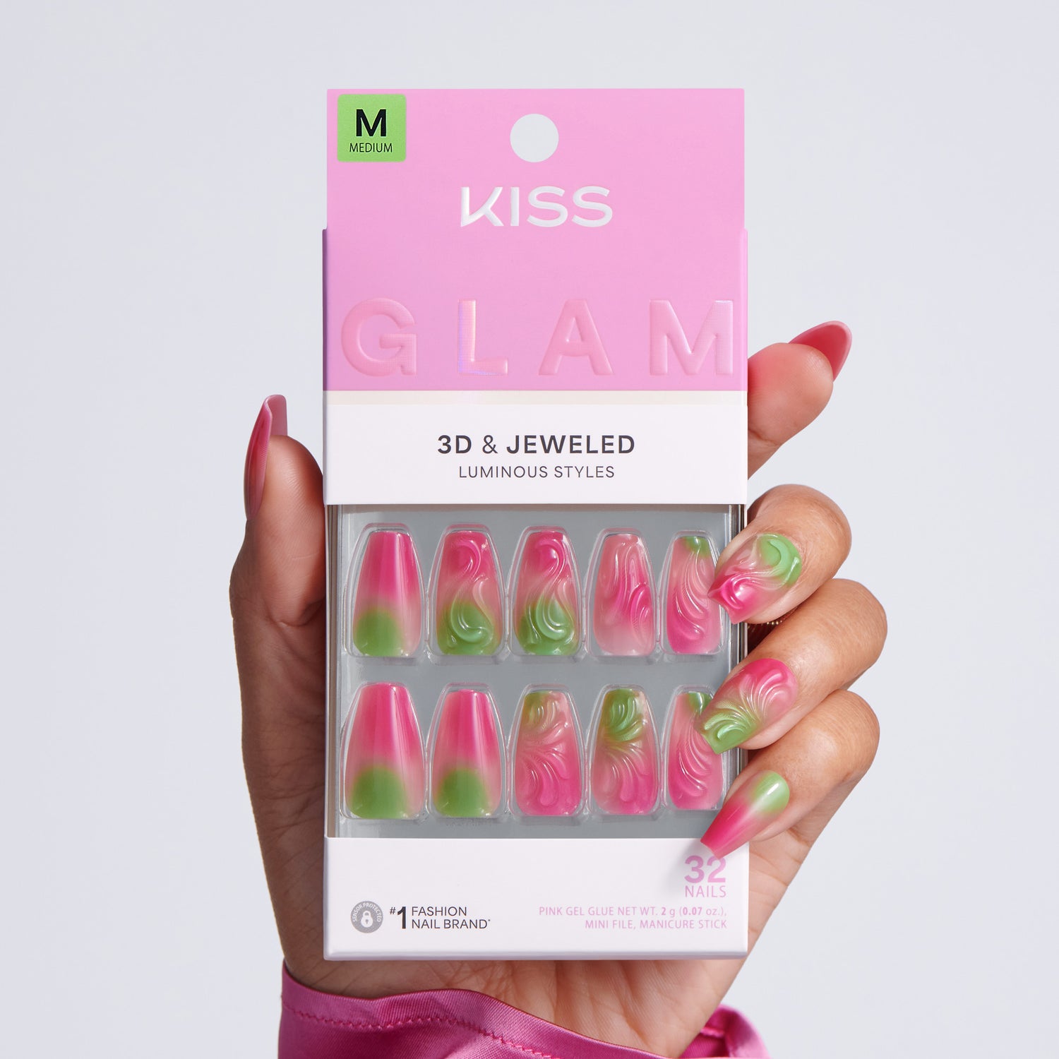 Hand holding KISS Glam 3D &amp; Jeweled Luminous Styles press-on nails packaging, Medium size, 32 nails, pink and green ombre coffin nails on light gray background
