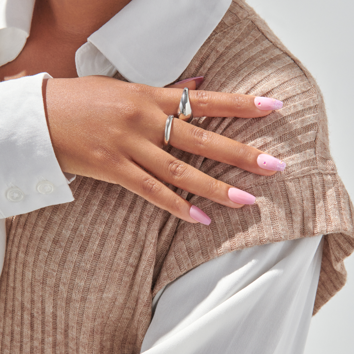 Hand with medium-length, square-shaped press-on nails in soft pink tones; some nails have purple glitter tips and small pink rhinestone accents.