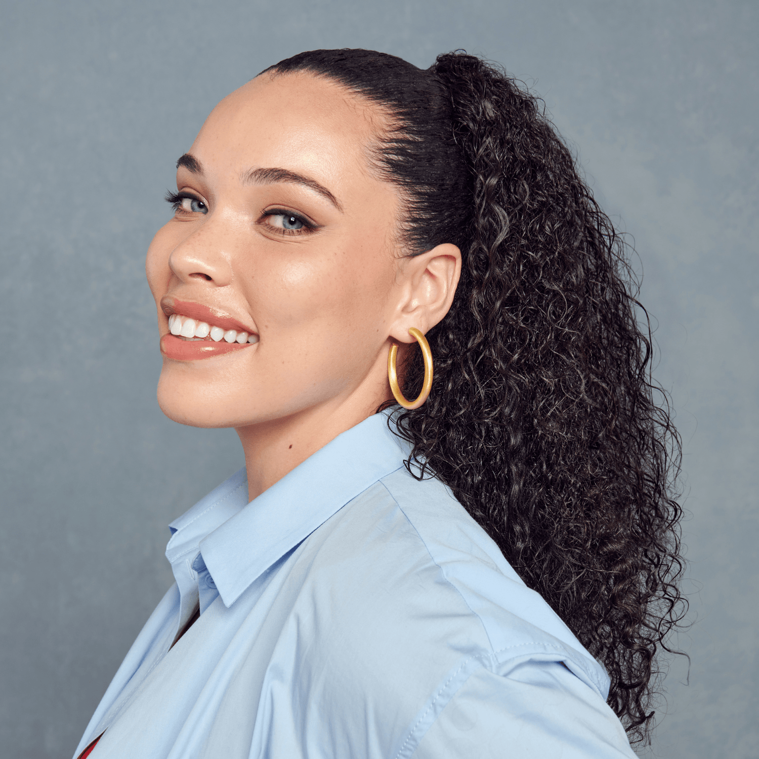 Woman with styled hair and gold hoop earrings against a gray background