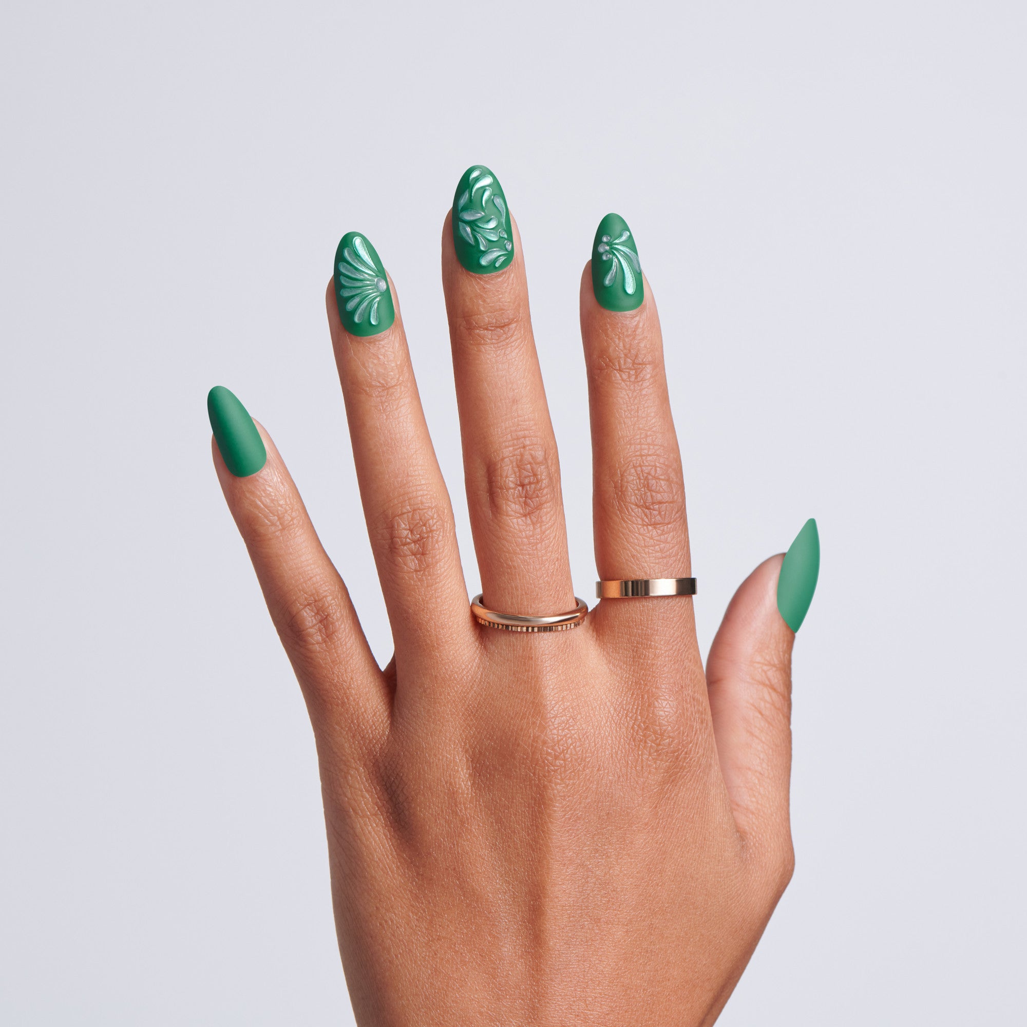 Hand with almond-shaped dark green press-on nails featuring white embossed floral nail art, gold ring, on light gray background