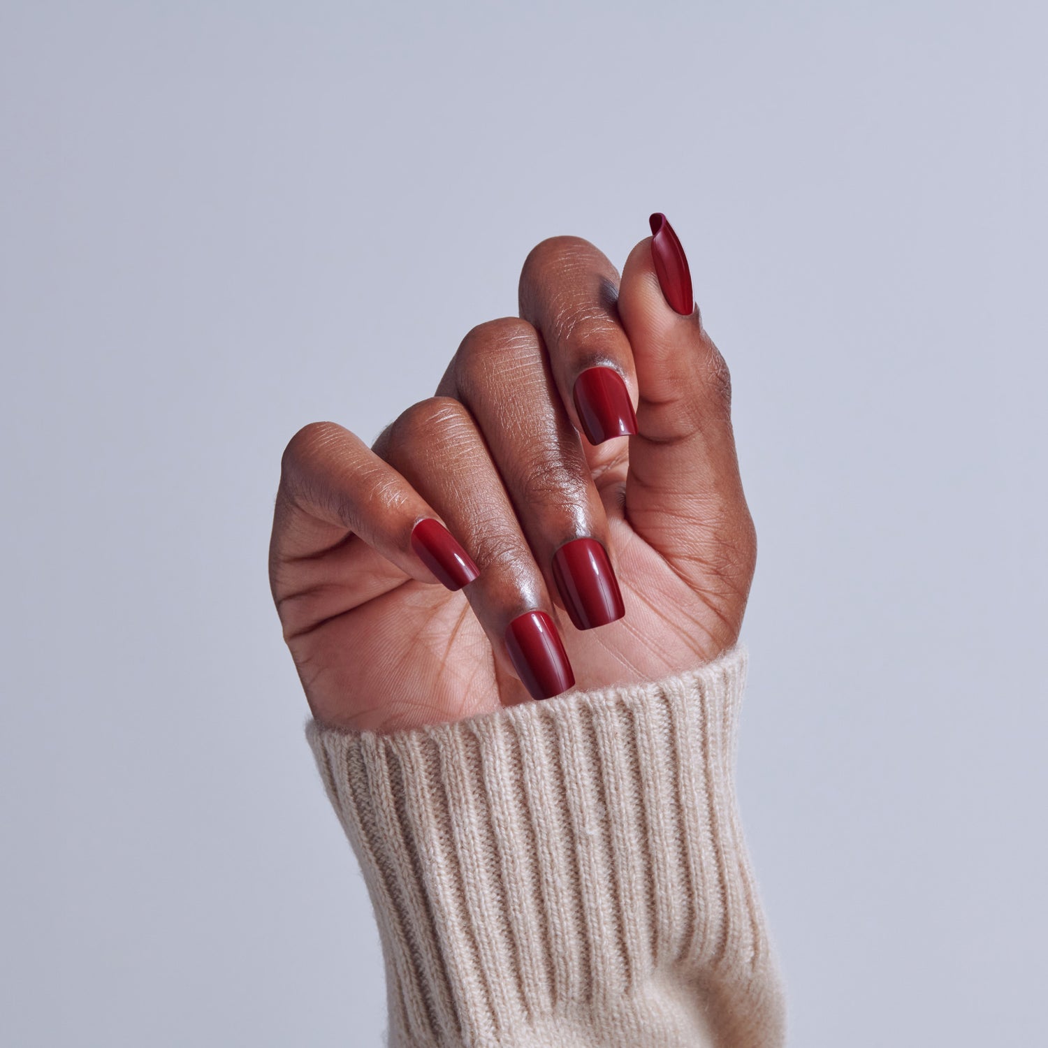Hand with deep red nail polish and almond-shaped nails wearing cream sweater against light background.
