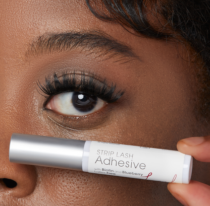 Close-up of a person holding a white and silver tube of strip lash adhesive near their eye with long, full lashes. The tube reads: \