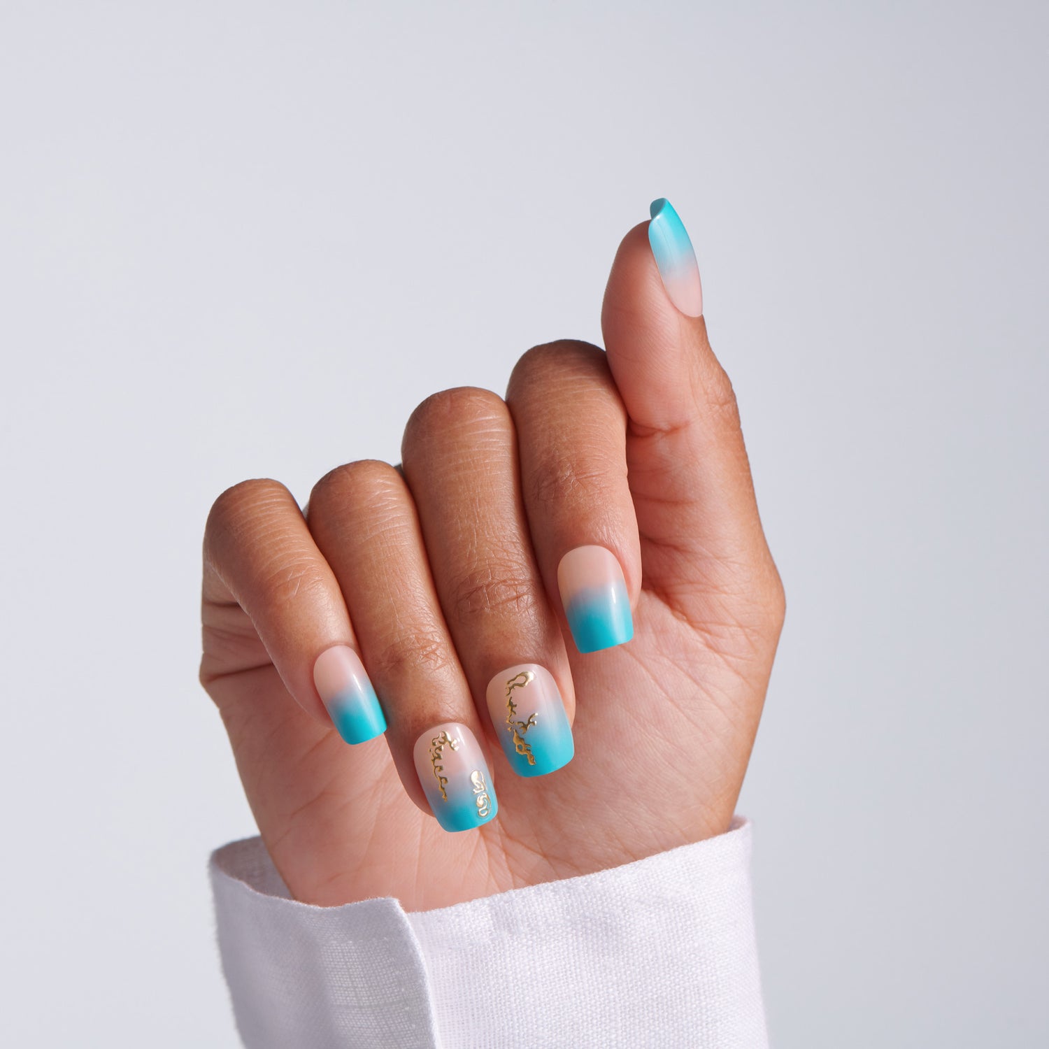 Hand with short squoval teal ombre press-on nails featuring gold chain accent nail art on light gray background