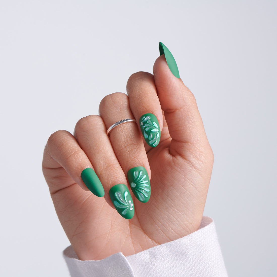 Close-up of hand with dark green almond-shaped press-on nails featuring white embossed floral designs, silver ring, on light gray background