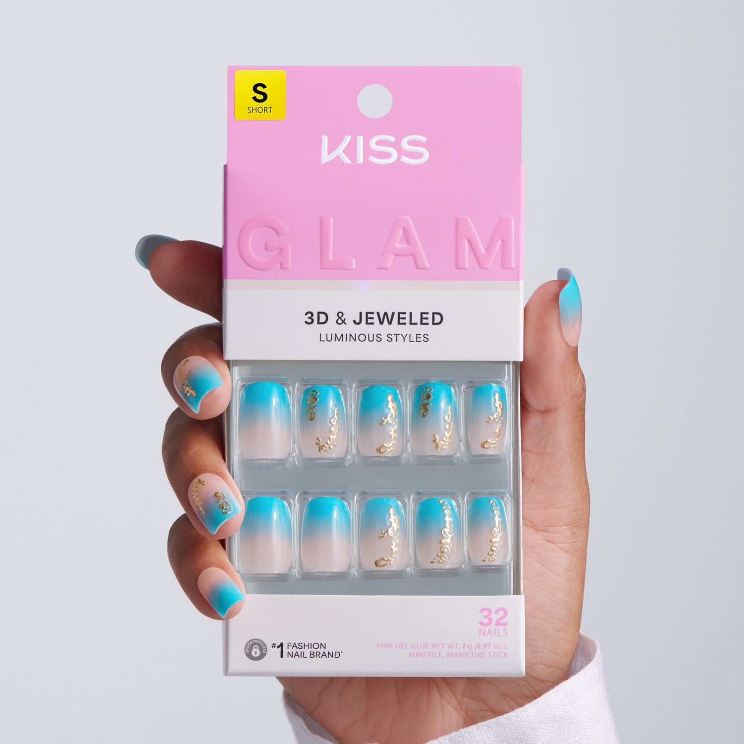 Hand holding KISS Glam 3D &amp; Jeweled Luminous Styles press-on nails packaging, Short size, 32 nails, teal ombre squoval nails with gold chain accents on light gray background