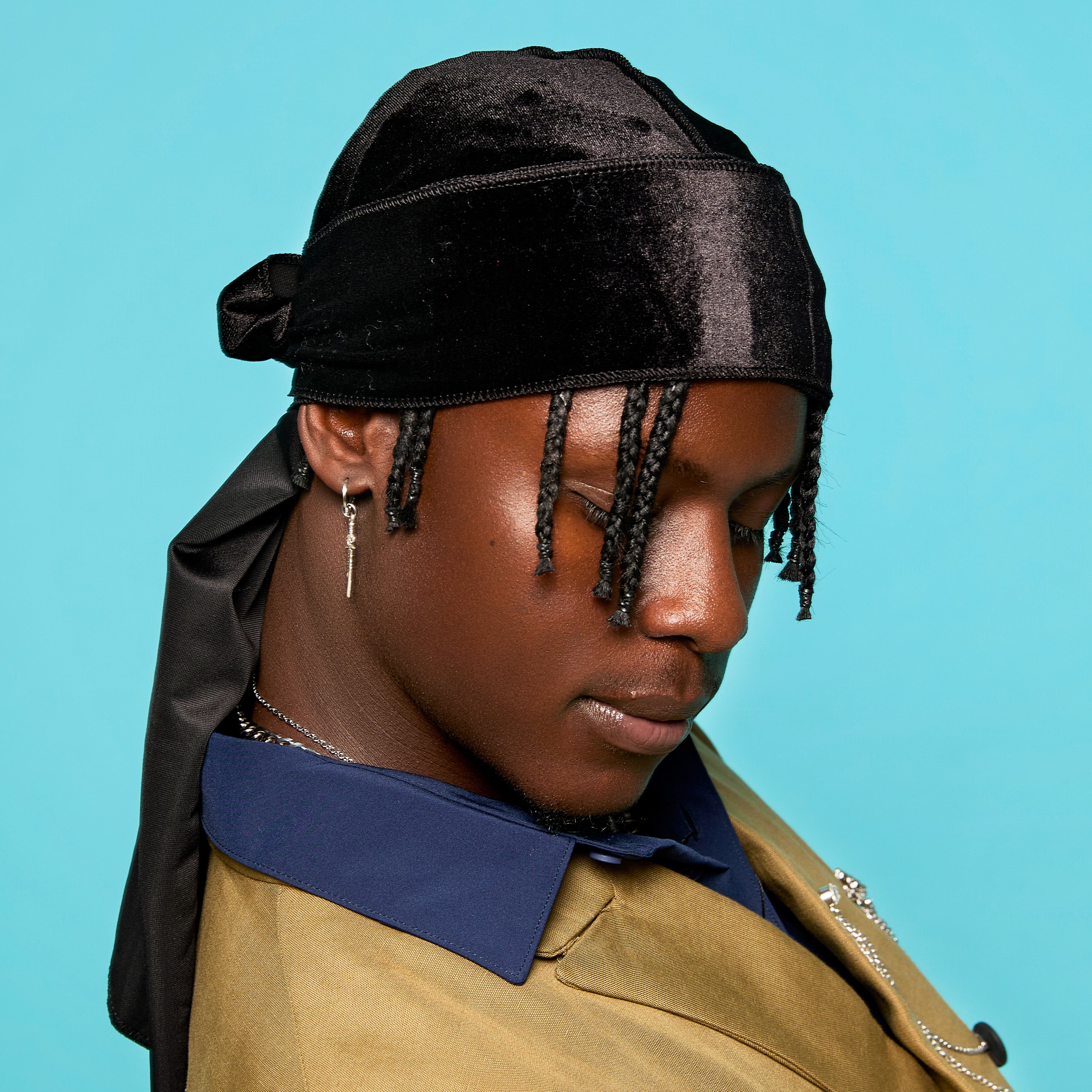 Black velvet durag tied at the back, worn by a person with braided hair and a blue and tan jacket, against a light blue background.