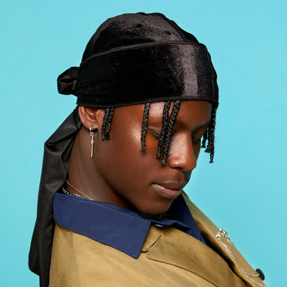 Black velvet durag tied at the back, worn by a person with braided hair and a blue and tan jacket, against a light blue background.