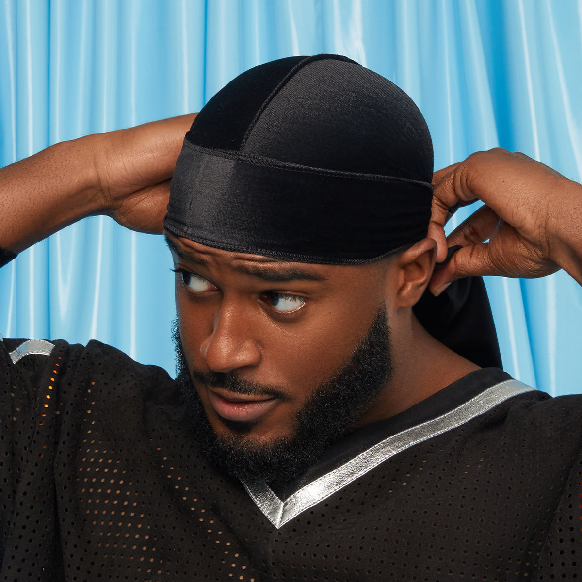 Black velvet durag worn by a man adjusting the back ties, against a blue background, highlighting a smooth, luxe fabric finish.