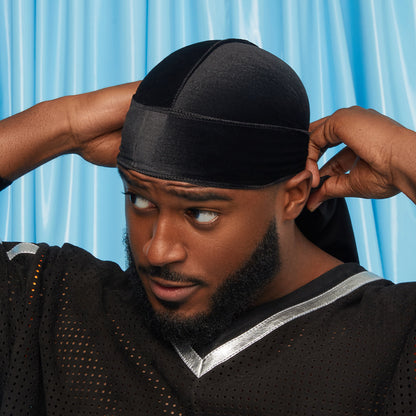 Black velvet durag worn by a man adjusting the back ties, against a blue background, highlighting a smooth, luxe fabric finish.
