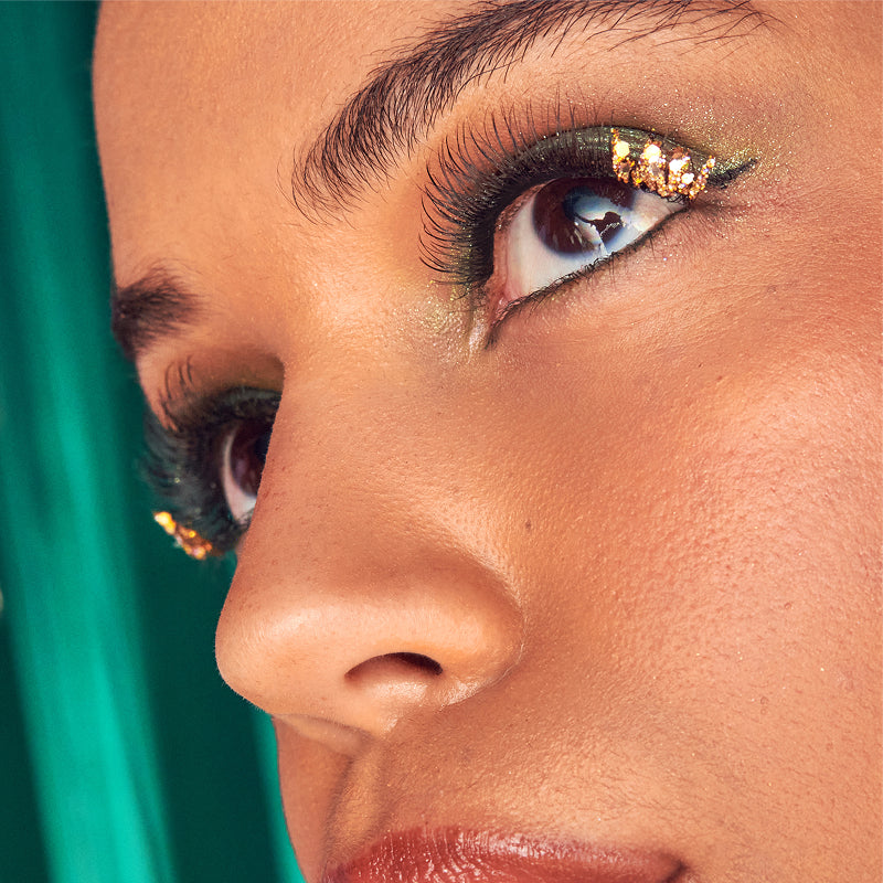 Close-up of model's face with gold glitter eyeshadow, winged eyeliner, and gold earrings against teal background.