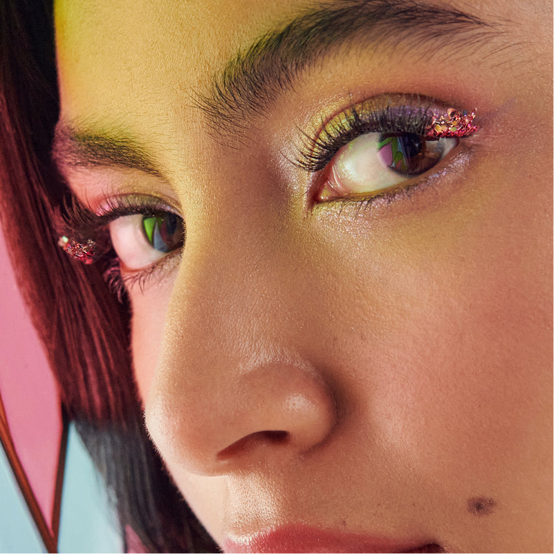 Close-up of model's face with colorful eyeshadow, glitter eyeliner, and burgundy hair against pink background.