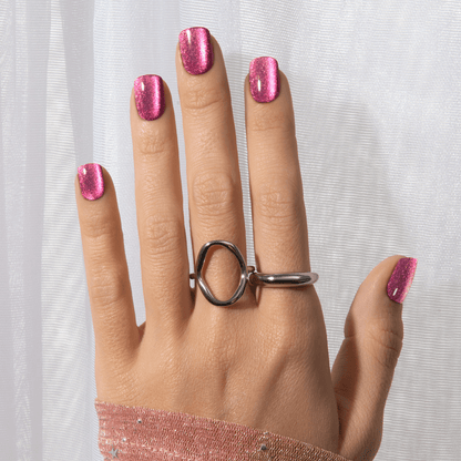 Hand with short, square-shaped press-on nails in glittery pink, wearing two silver rings, displayed against a sheer white curtain background.
