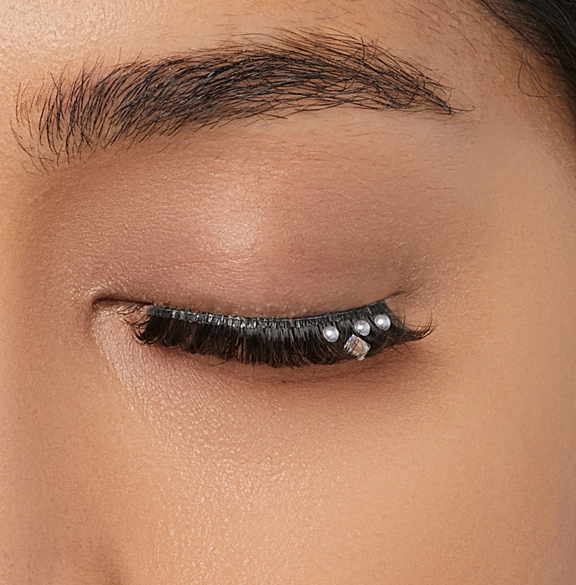 Closed eye with dark, spiky false eyelashes adorned with three small pearls and one square jewel near the outer corner.