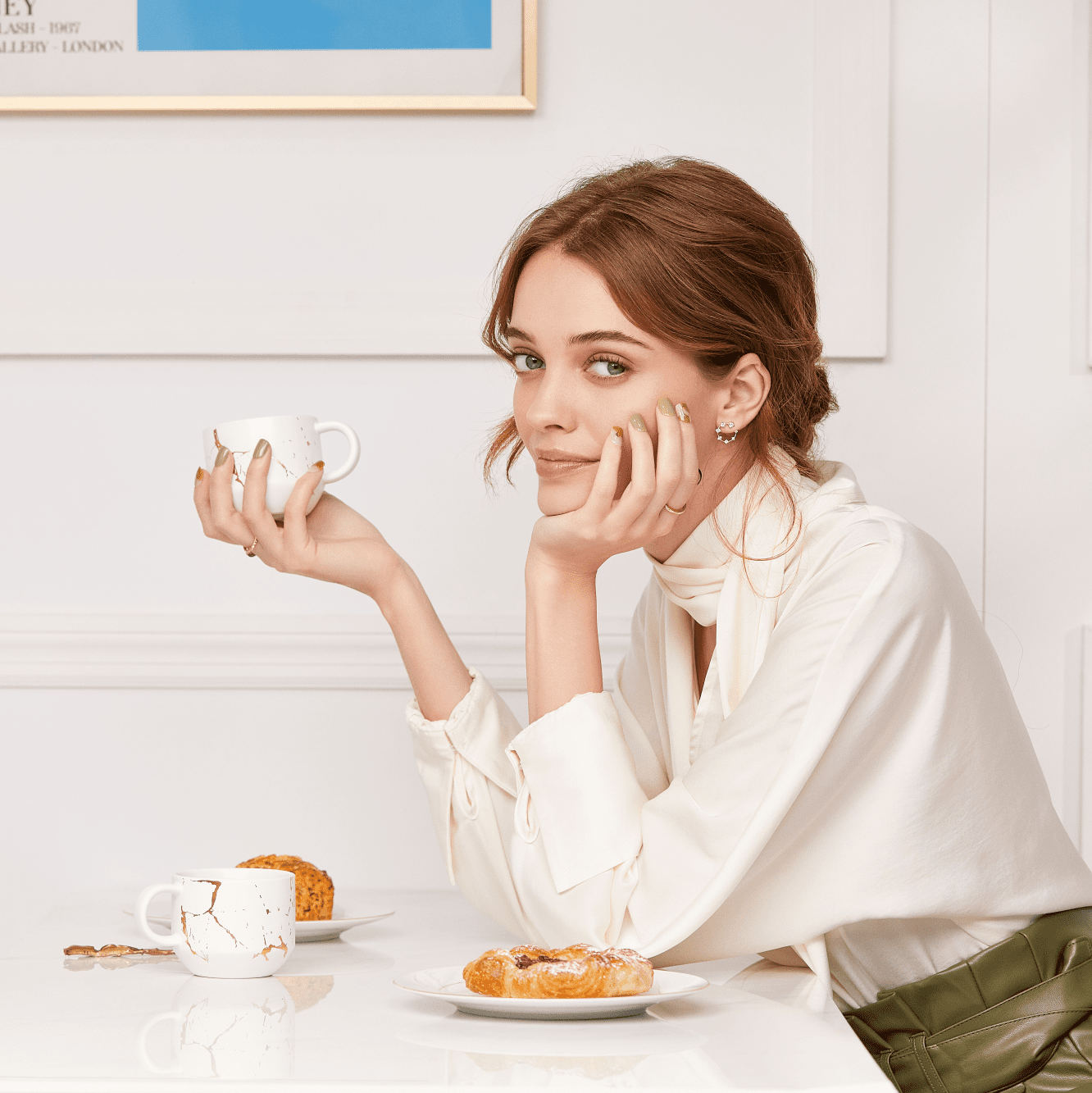 Young woman sitting at a white table holding a white marbled teacup, wearing olive green and beige press-on nails with gold accents, dressed in a cream blouse.