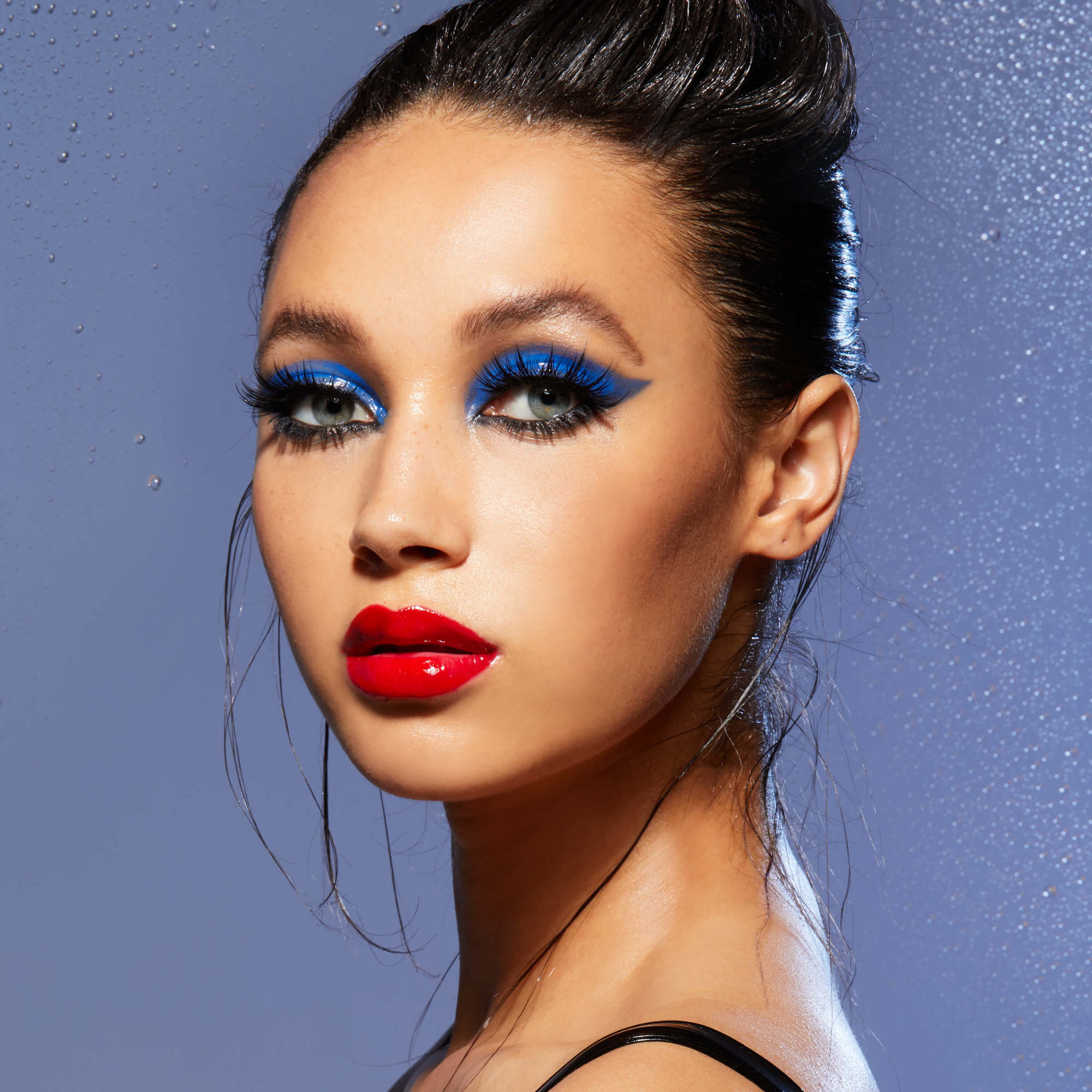 Woman with bold blue eyeshadow, red lipstick, and slicked-back hair against sparkly blue background.