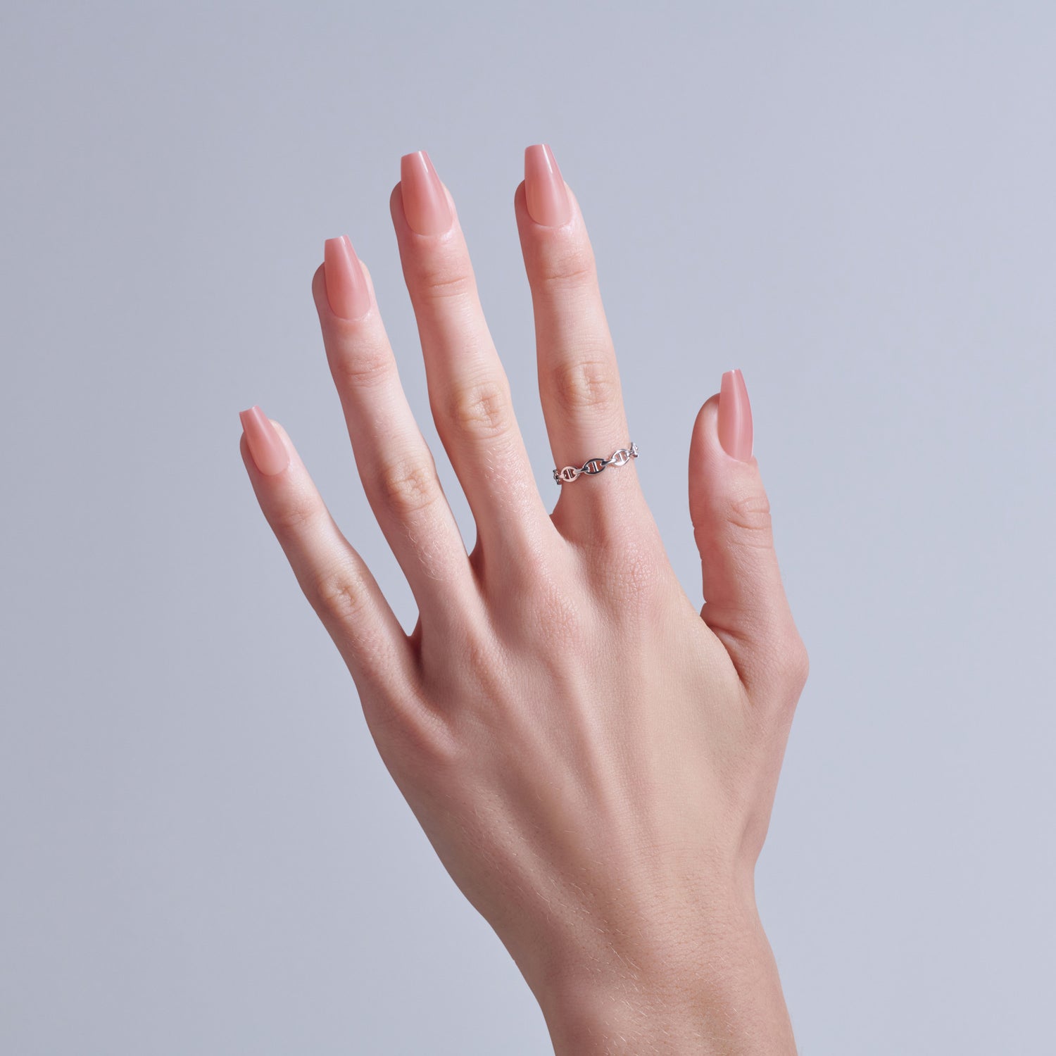 Hand with elegant coffin-shaped nails and silver chain ring against gray background.