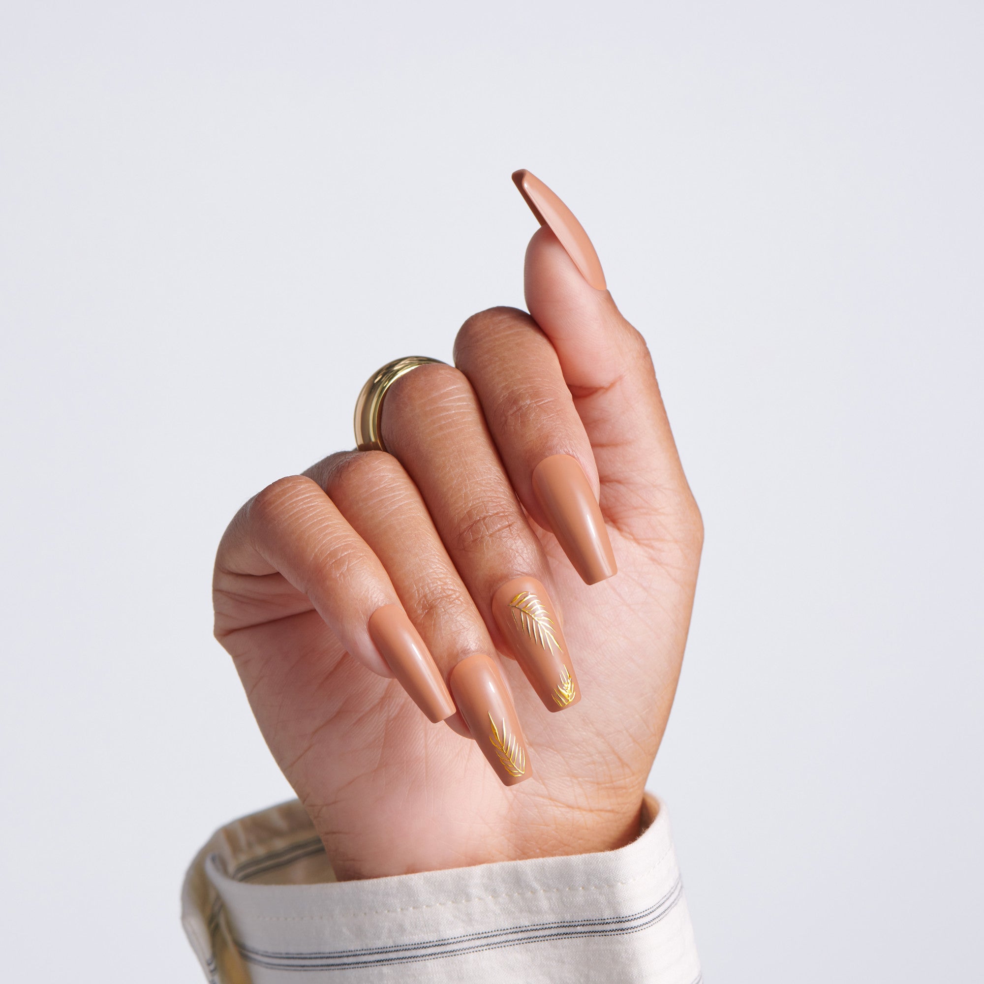 Hand with long coffin-shaped nude nails and a gold leaf accent design on one nail, wearing a gold ring, light gray background