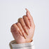 Hand with long coffin-shaped nude nails and a gold leaf accent design on one nail, wearing a gold ring, light gray background