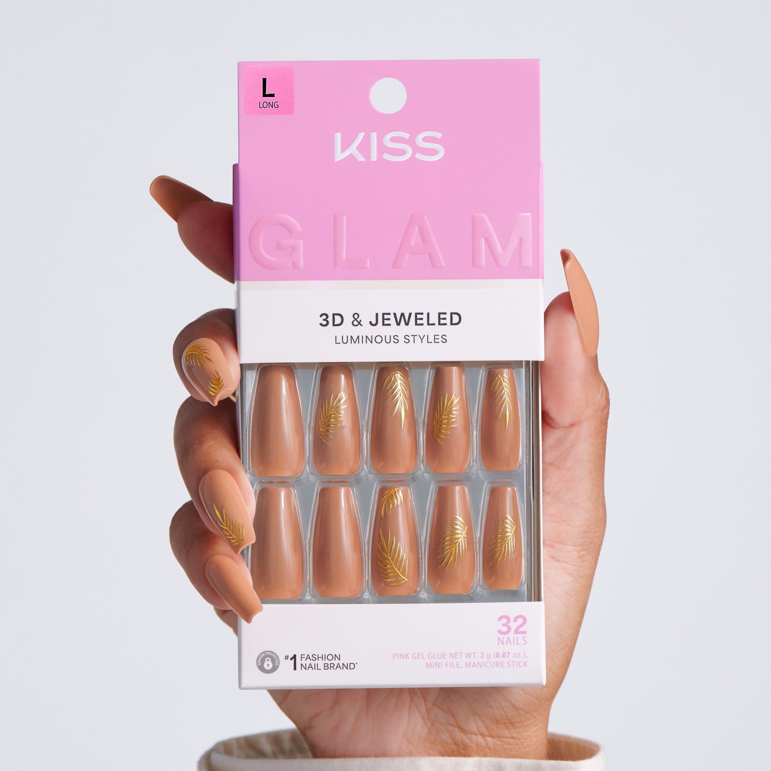 Hand holding KISS Glam 3D &amp; Jeweled Luminous Styles press-on nails packaging, Long size, 32 nails, pink box showing nude nails with gold leaf accents through clear window