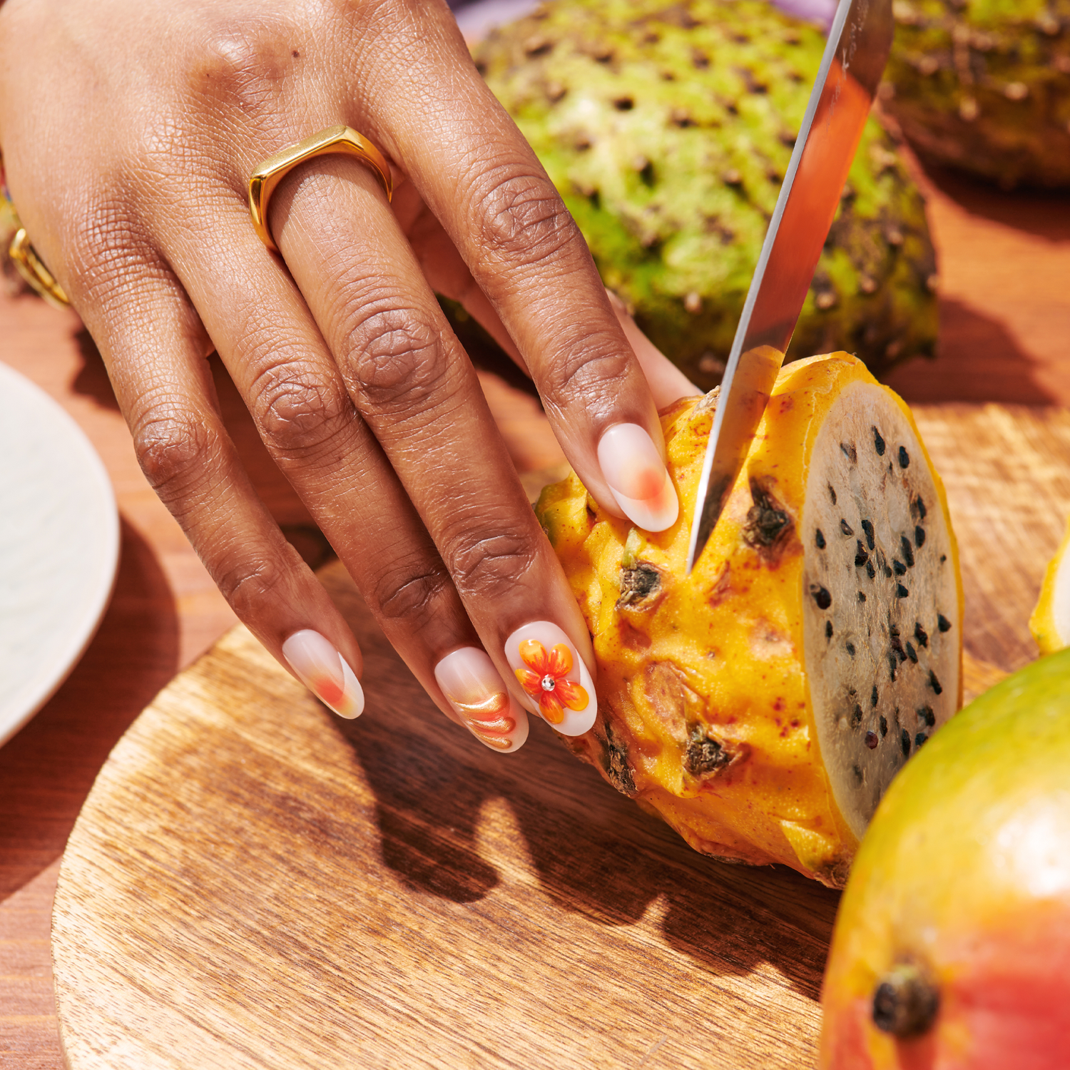 Hand with oval-shaped press-on nails featuring white base with orange floral nail art accents, gold ring on finger, holding sliced dragon fruit on a wooden cutting board