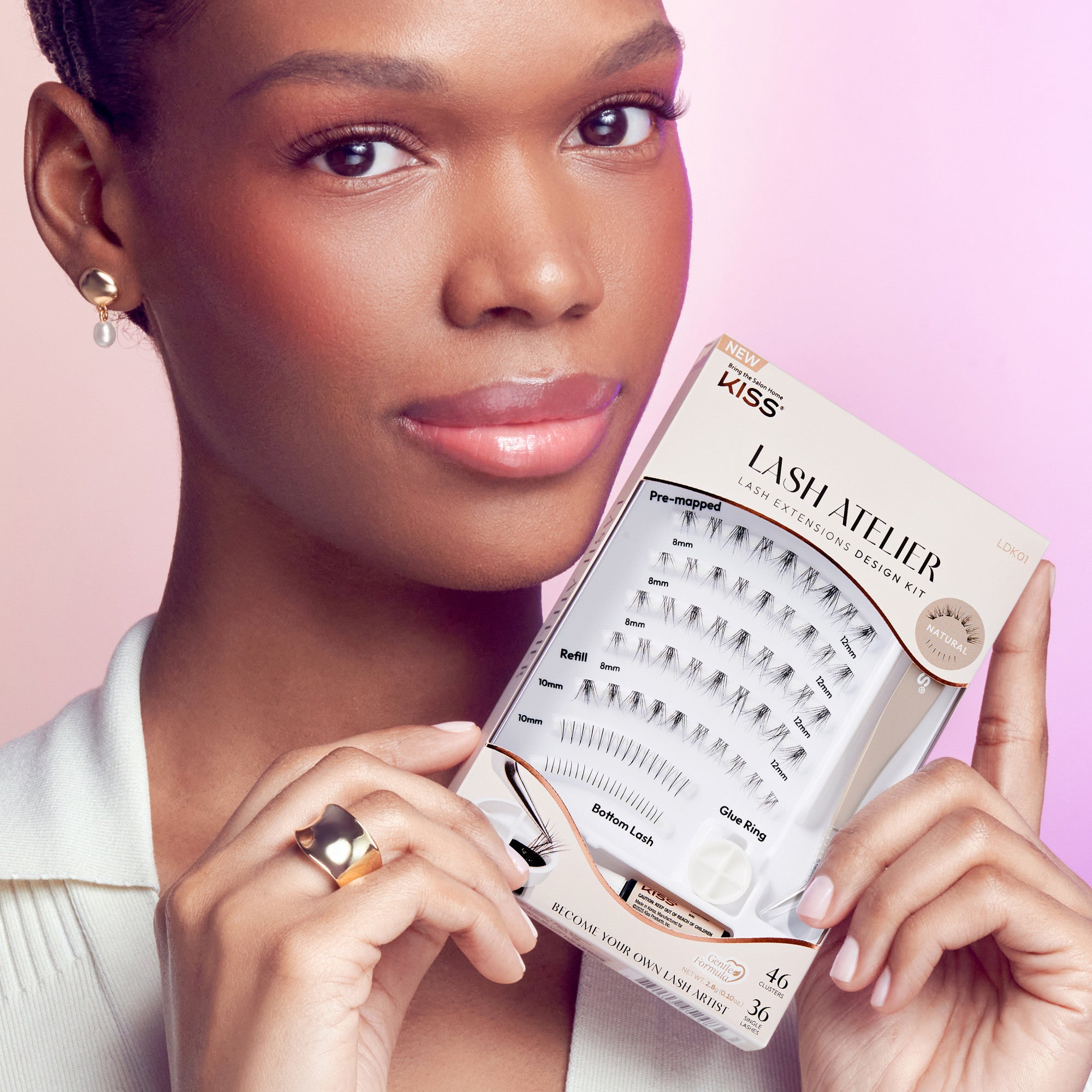 Close-up of a woman with natural makeup holding the KISS Lash Atelier Individual Lash Extensions Design Kit, showing pre-mapped and refill lash clusters in various lengths, a glue ring, and lash glue, labeled \