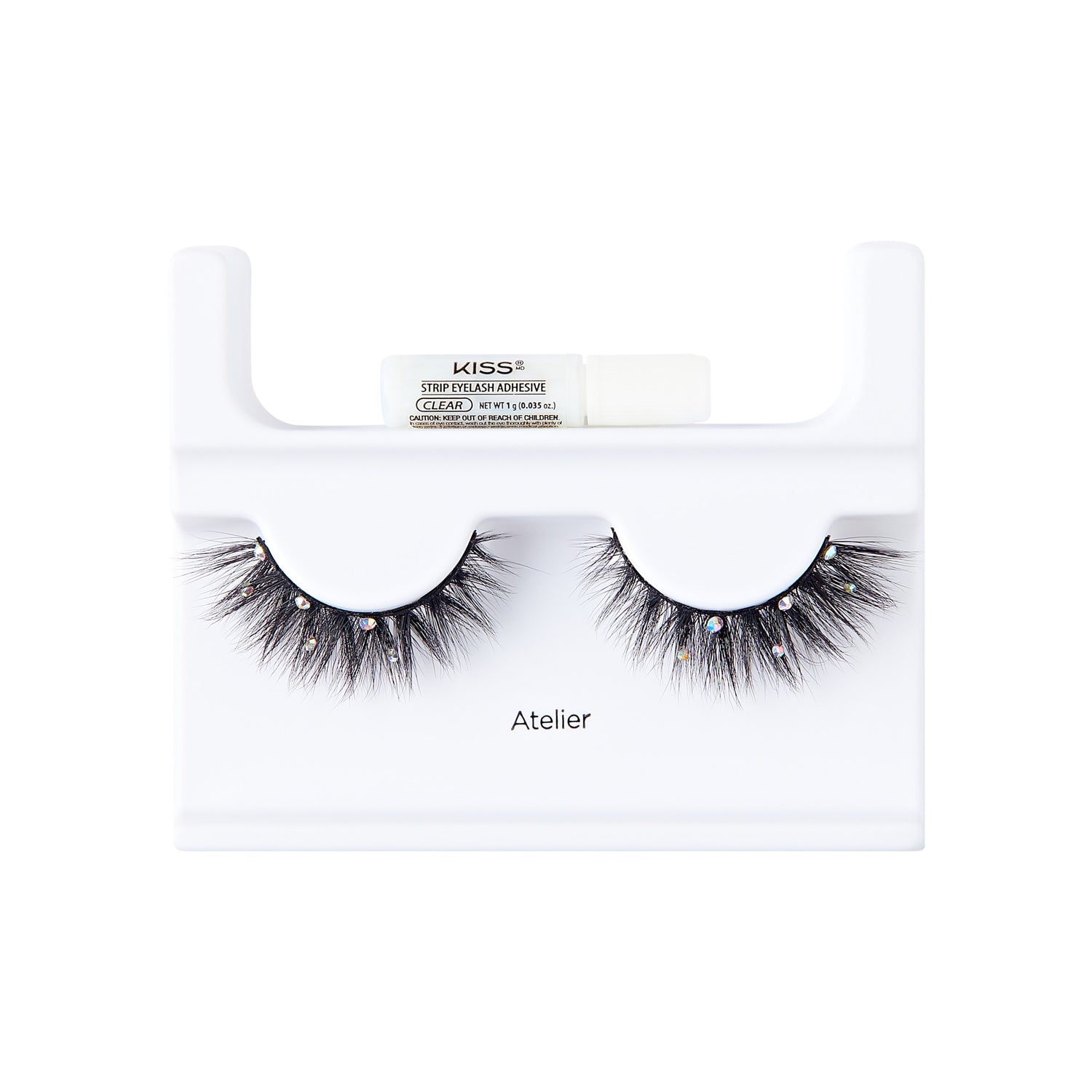 The image shows a pair of false eyelashes displayed on a white plastic holder. The eyelashes are thick, voluminous, and have a dramatic flair with small decorative elements, possibly rhinestones, attached to them. Above the eyelashes, there is a small tube of adhesive labeled "KISS" with additional text indicating it is a strip lash adhesive. The word "Atelier" is printed on the holder below the eyelashes. The overall presentation is clean and professional, suitable for retail display.