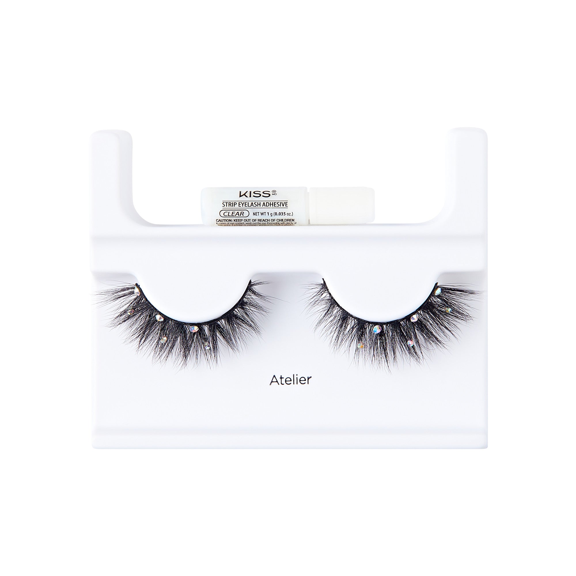 The image shows a pair of false eyelashes displayed on a white plastic holder. The eyelashes are thick, voluminous, and have a dramatic flair with small decorative elements, possibly rhinestones, attached to them. Above the eyelashes, there is a small tube of adhesive labeled "KISS" with additional text indicating it is a strip lash adhesive. The word "Atelier" is printed on the holder below the eyelashes. The overall presentation is clean and professional, suitable for retail display.