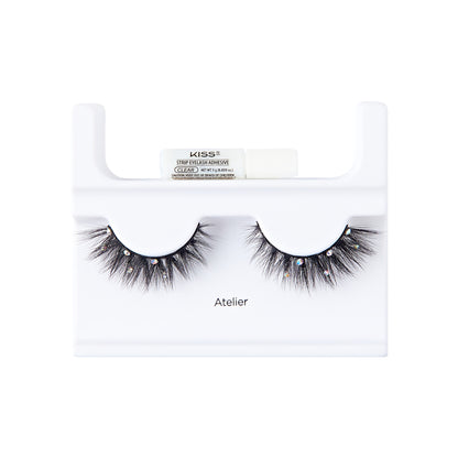 The image shows a pair of false eyelashes displayed on a white plastic holder. The eyelashes are thick, voluminous, and have a dramatic flair with small decorative elements, possibly rhinestones, attached to them. Above the eyelashes, there is a small tube of adhesive labeled "KISS" with additional text indicating it is a strip lash adhesive. The word "Atelier" is printed on the holder below the eyelashes. The overall presentation is clean and professional, suitable for retail display.