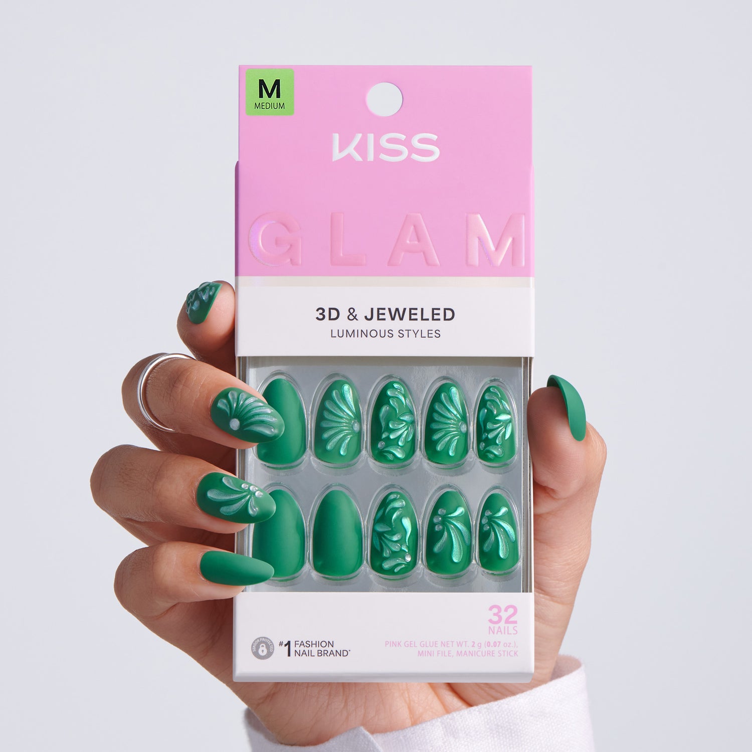 Hand holding KISS Glam 3D &amp; Jeweled Luminous Styles press-on nails packaging, Medium size, 32 nails, dark green almond nails with white embossed floral designs on light gray background