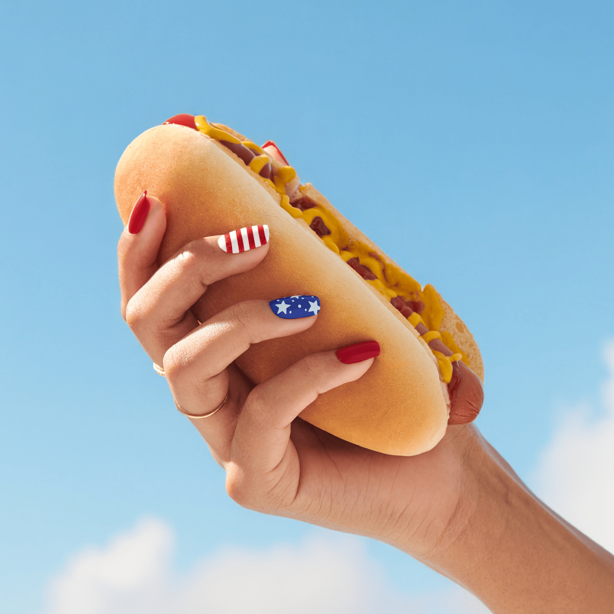 Hand holding a hot dog with mustard against a clear blue sky