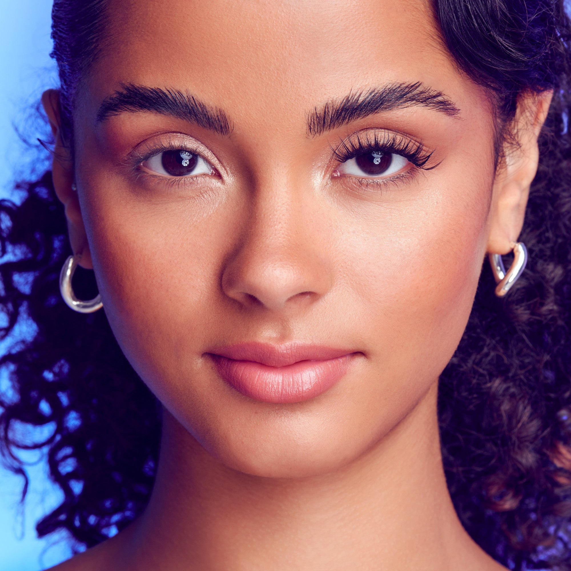 The image features a close-up portrait of a young woman with curly hair. She has a warm complexion and is wearing subtle makeup that highlights her natural beauty. Her eyebrows are well-defined, and her eyes are expressive, showcasing a soft gaze. She is adorned with silver hoop earrings, adding a touch of elegance to her look. The background is softly lit, creating a pleasing contrast that enhances her features.