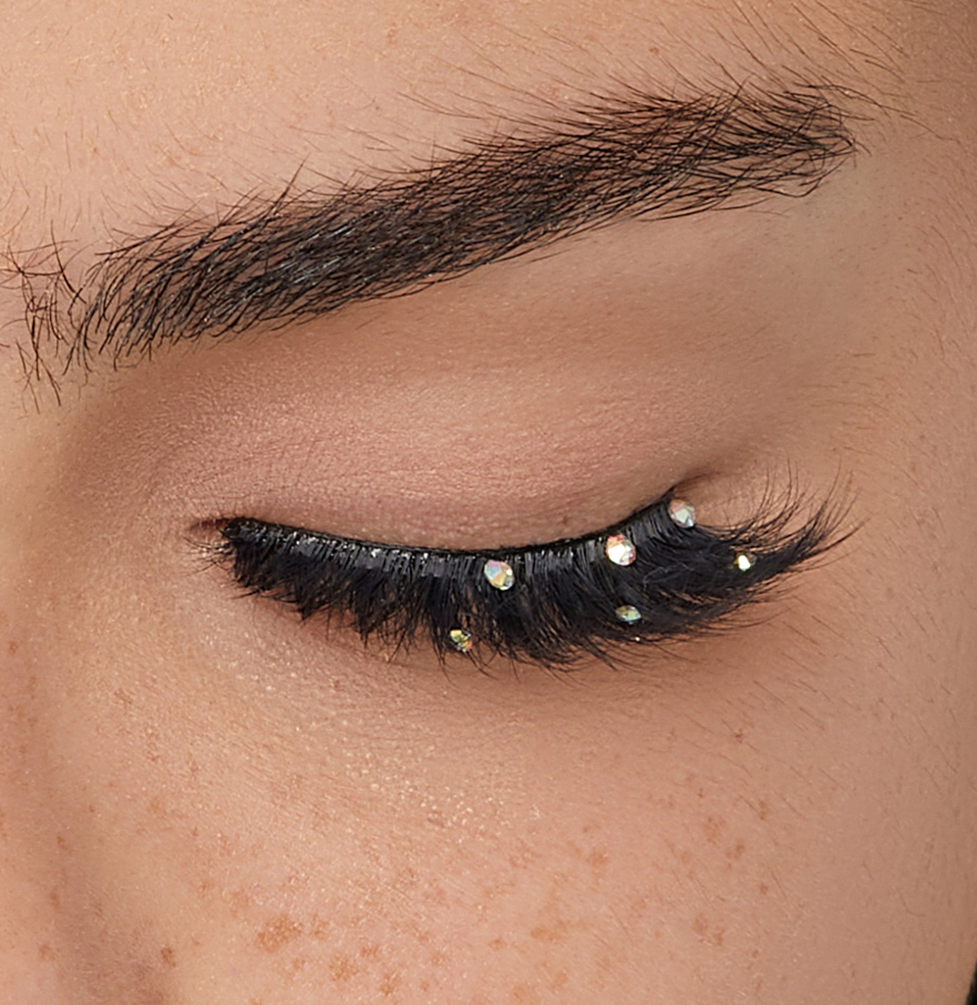 The image shows a close-up of a closed eye. The person has thick, well-groomed eyebrows and long, dark eyelashes. The eyelashes are adorned with small, shiny rhinestones, adding a decorative and glamorous touch. The skin around the eye is smooth, with a few freckles visible. The overall look is elegant and stylish.