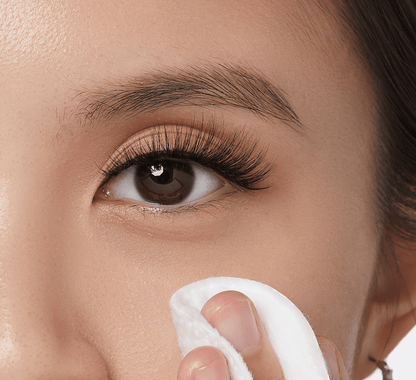 Close-up of a brown eye with long, curled lashes and shimmery nude eyeshadow, as a hand gently wipes the skin near the eye with a white cotton pad.