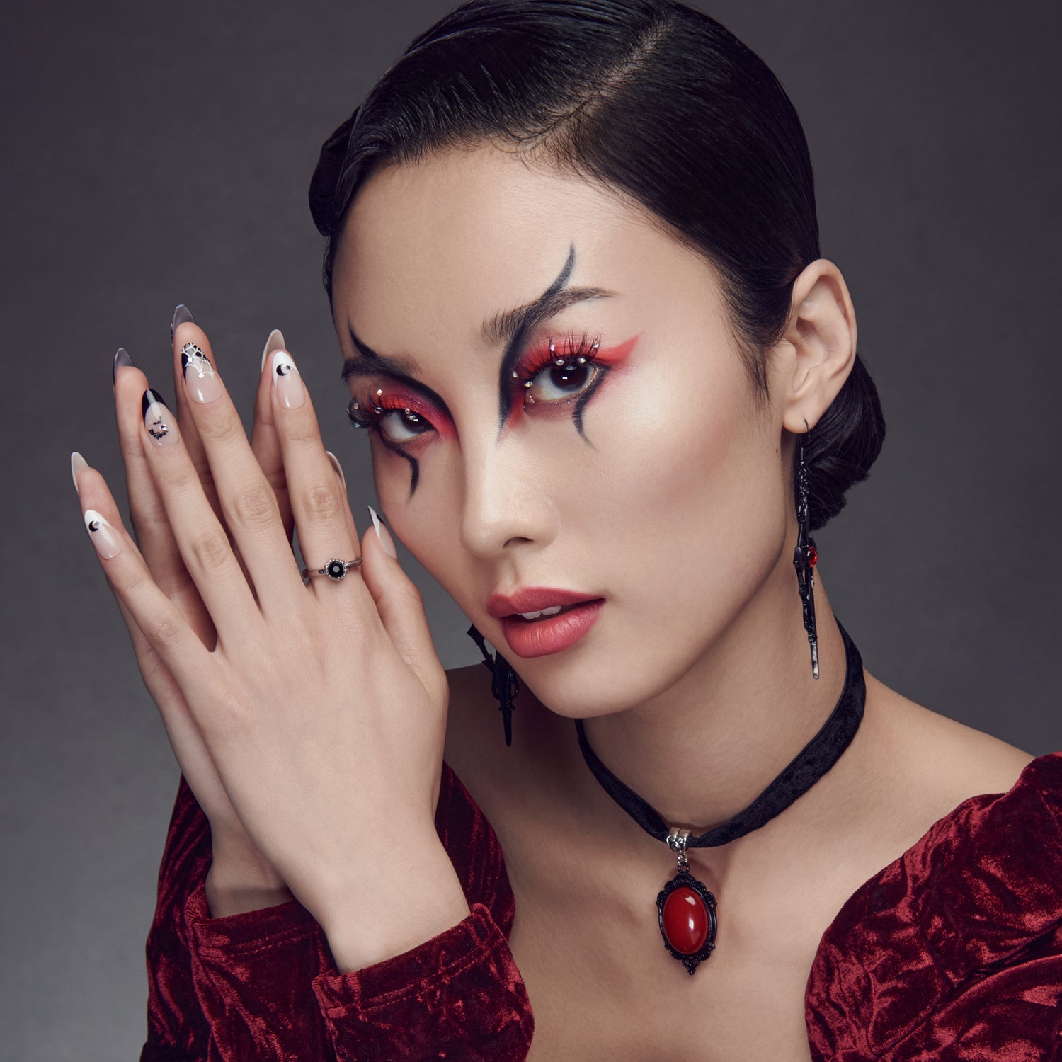 Close-up of a woman wearing decorative false eyelashes with black and red spikes, bold red smoky eye makeup, moon-inspired nail art, a velvet red top, and matching gothic jewelry.