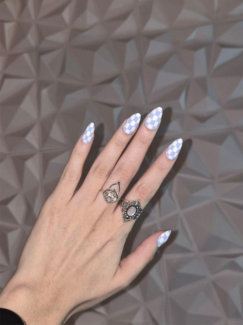 Almond nails with periwinkle gingham pattern and ornate silver rings on textured background.