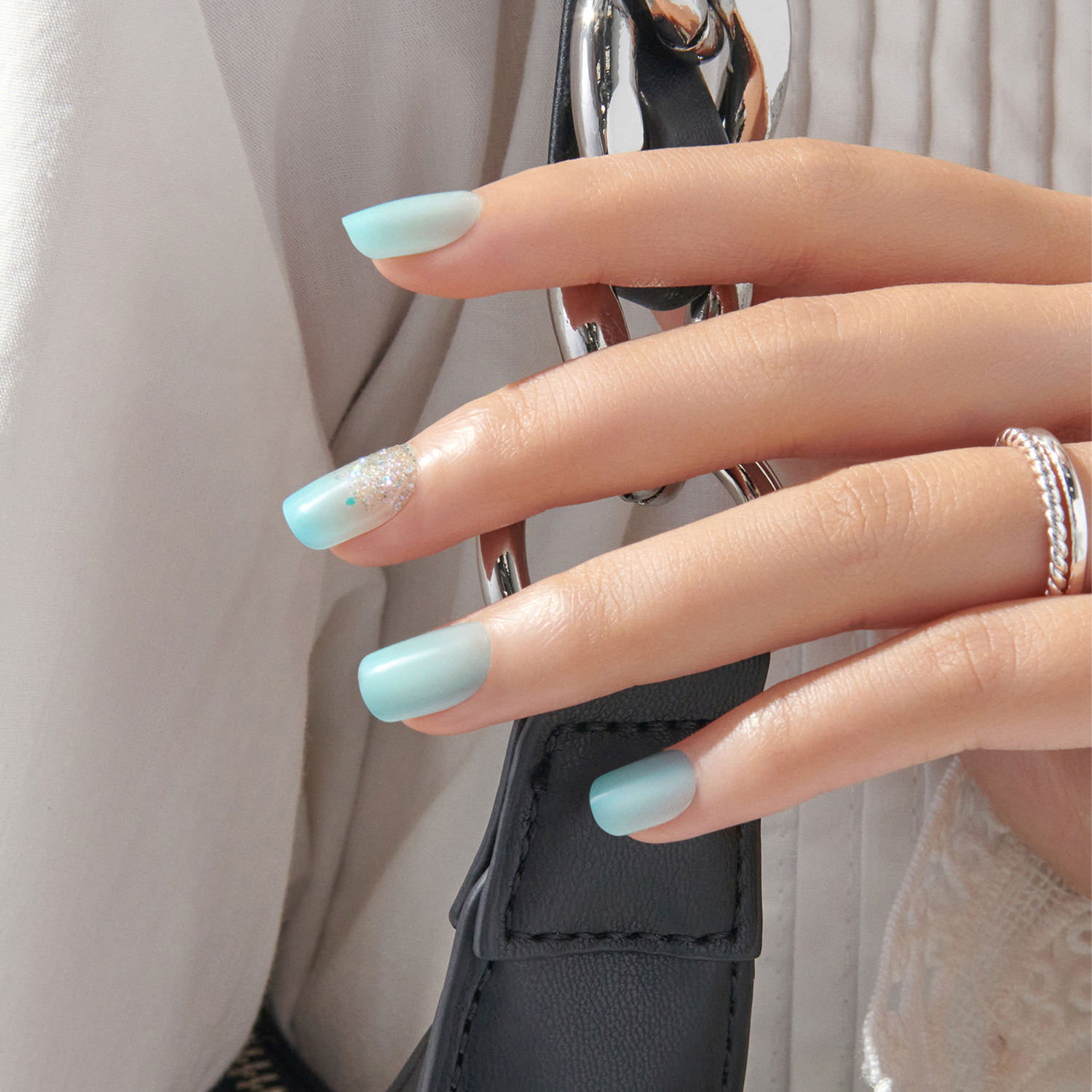 Hand with light blue matte press-on nails, one accent nail featuring silver glitter, holding a black leather strap with silver chain detail.