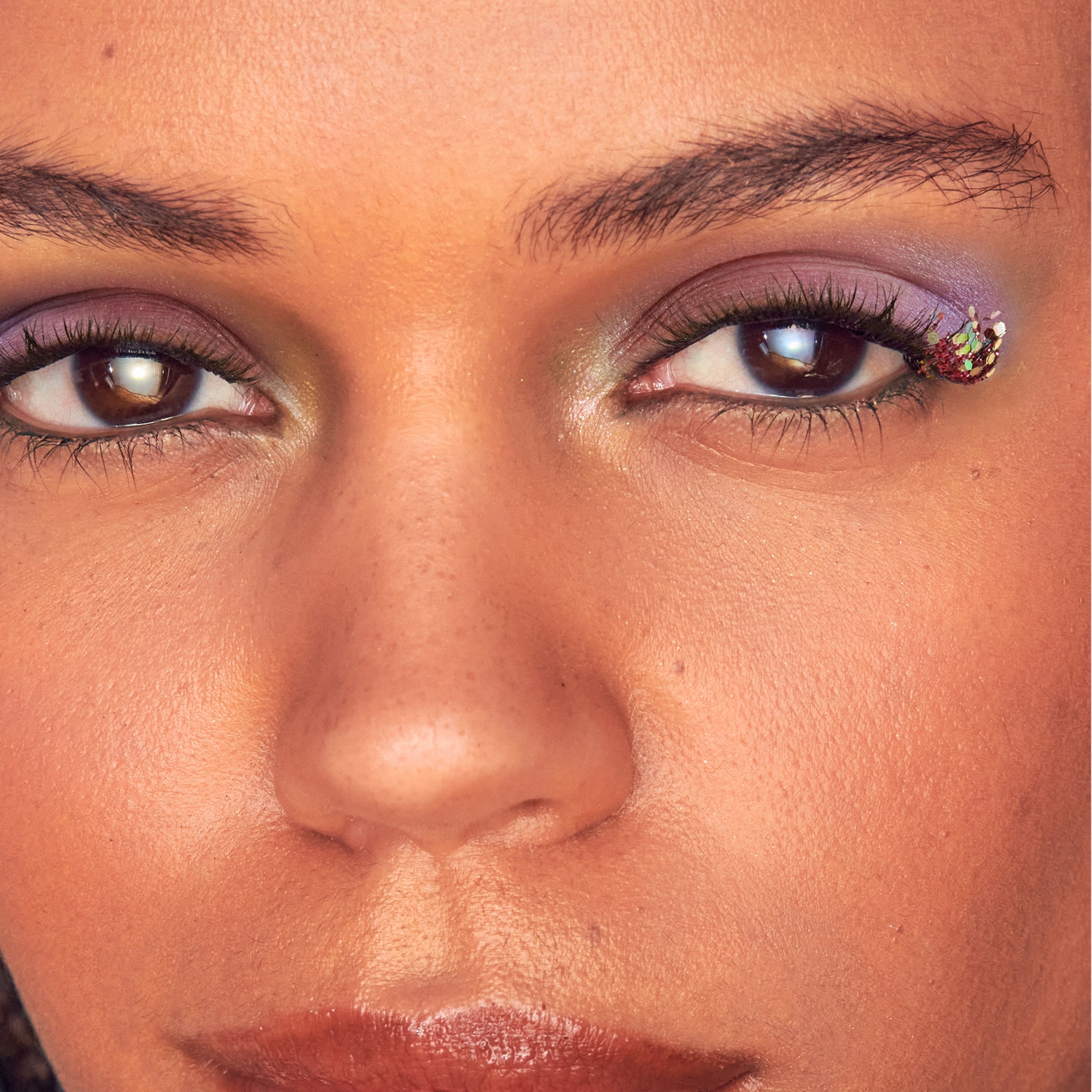 Close-up of a person’s eyes with purple eyeshadow and a decorative cluster of iridescent glitter and sequins near the outer corner of the right eye.