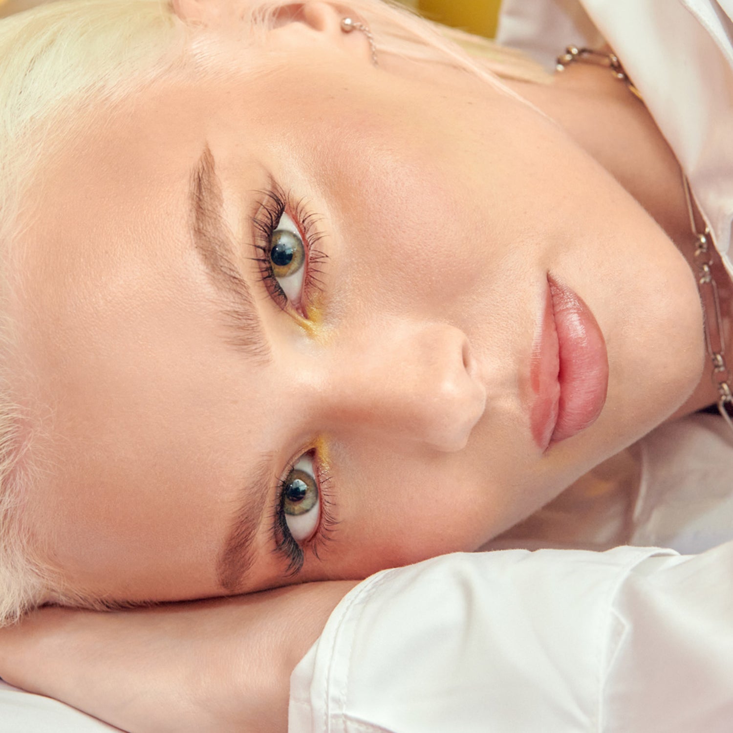 Close-up of a person with platinum blonde hair wearing long, self-adhesive false eyelashes and subtle yellow eyeshadow, resting their head on their arm.