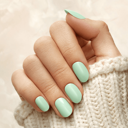 Hand with press-on nails in a soft mint green color, shown against a light cream knit sweater background, nails are oval shaped and glossy.