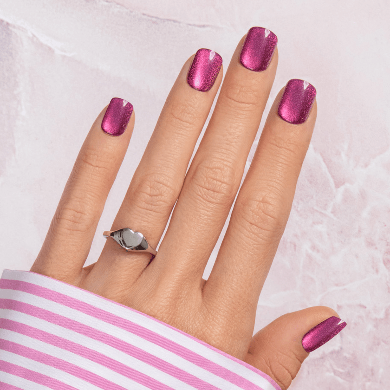 Hand wearing pink and purple metallic press-on nails in a medium square shape, with a silver heart-shaped ring and a pink striped shirt cuff.