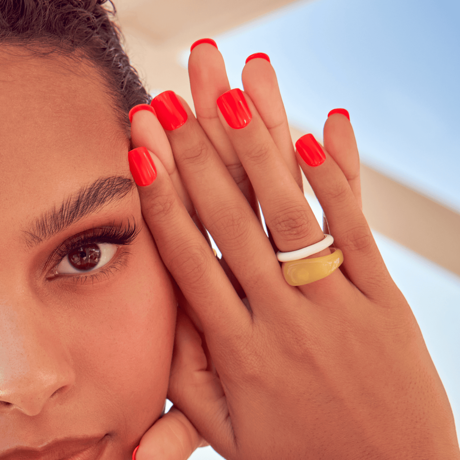 Close-up of a person’s hand with bright coral red square-shaped press-on nails, wearing chunky white and yellow rings, against a soft blue and beige background.