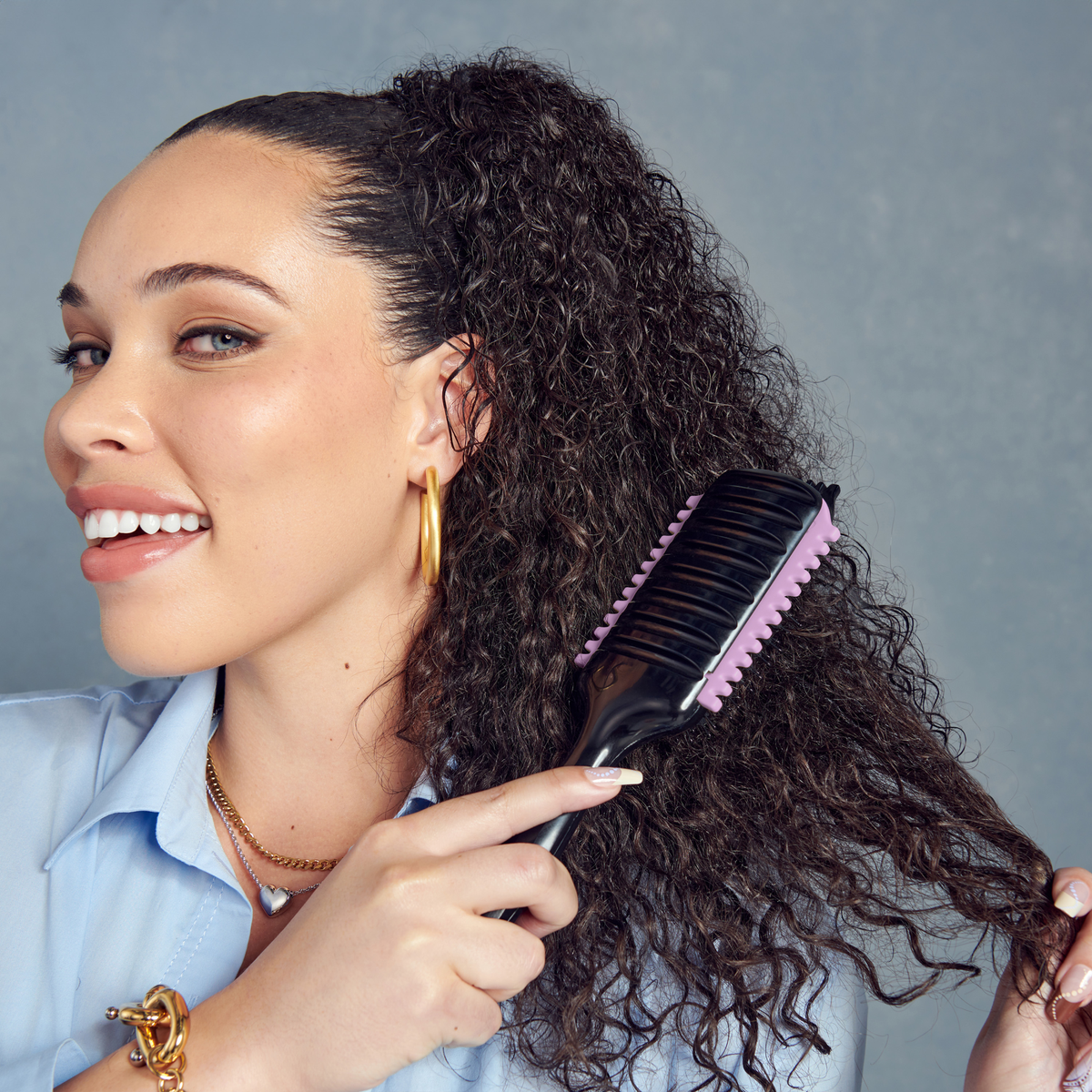 Woman with curly dark hair smiling while using a black and lavender KISS Colors &amp; Care SwitchCurl™ Define Brush on her hair against a gray background.
