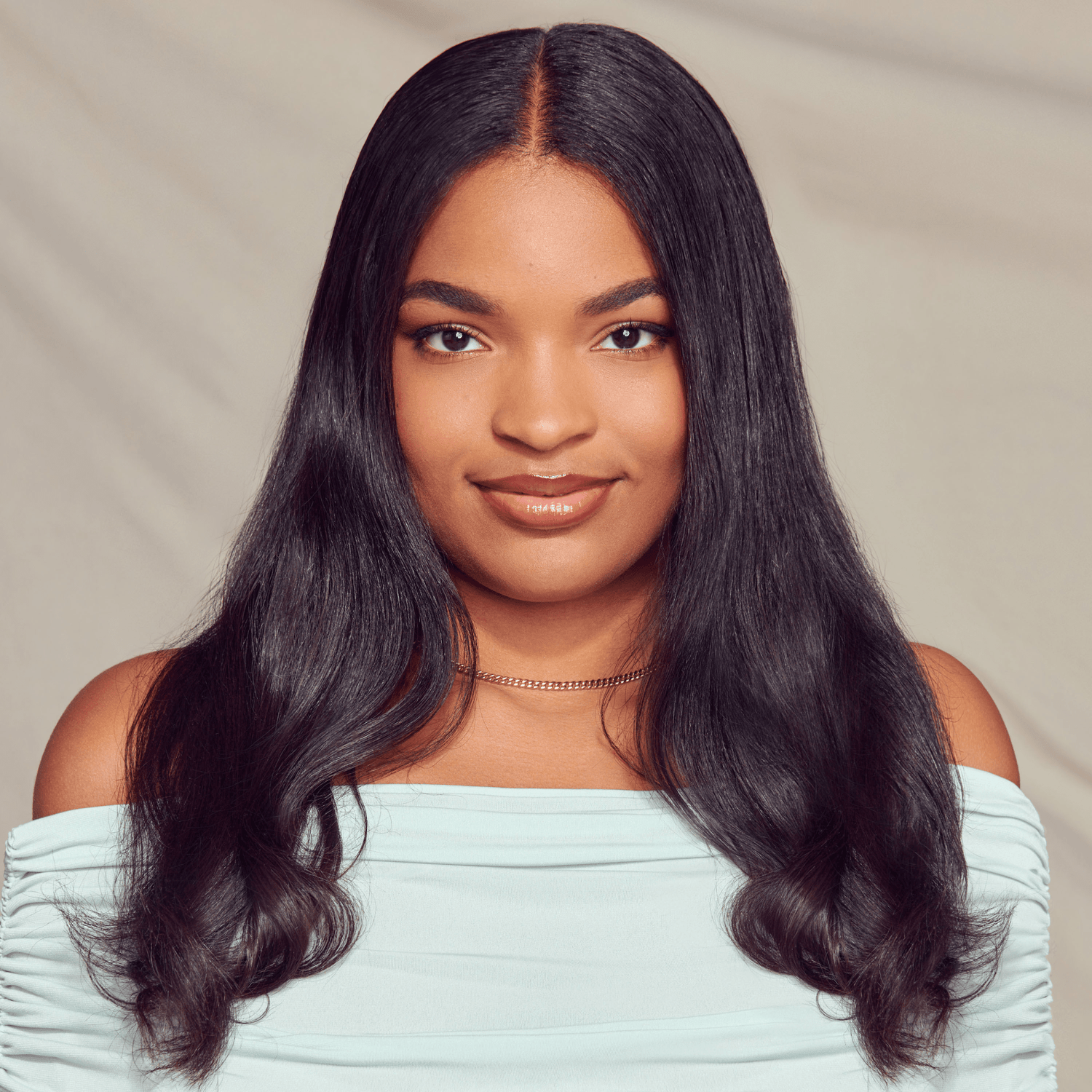 Young woman with long, smooth, shiny black hair wearing a light blue off-shoulder top and a delicate necklace, smiling softly against a neutral background.