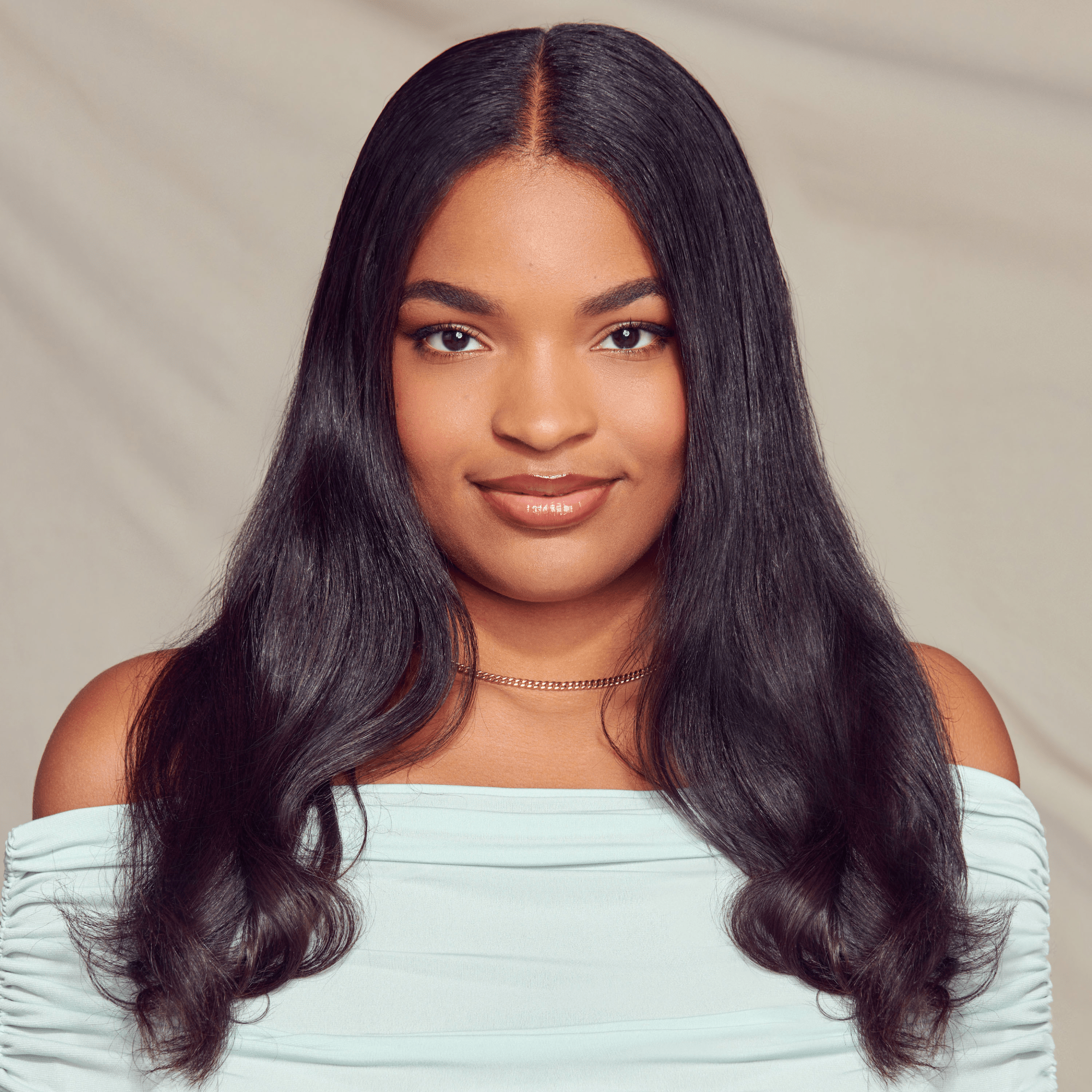 Young woman with long, smooth, shiny black hair wearing a light blue off-shoulder top and a delicate necklace, smiling softly against a neutral background.