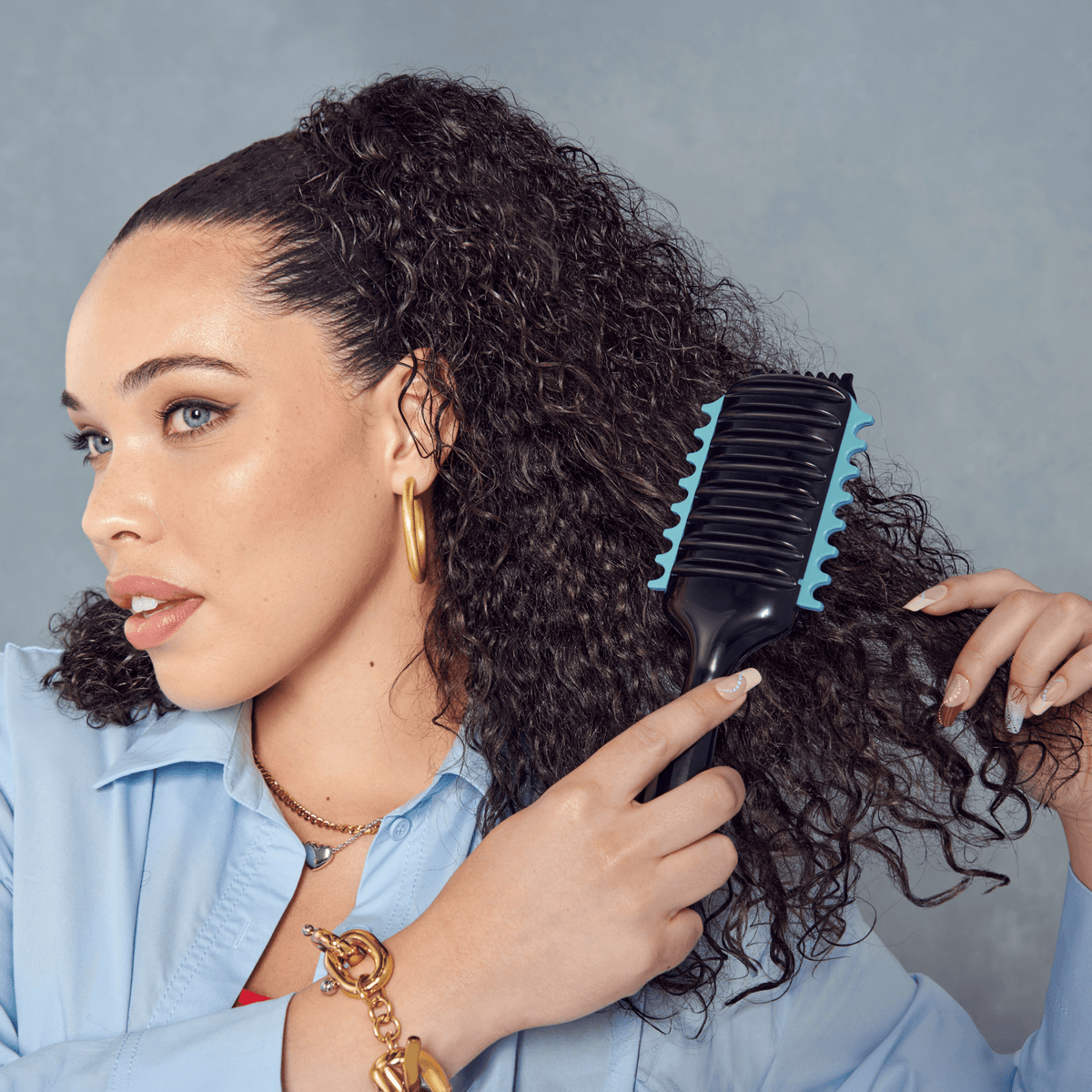 Woman with curly dark hair using a black and blue KISS SwitchCurl™ Define Brush, wearing gold jewelry and a light blue shirt against a gray background.
