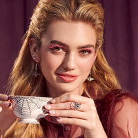 Woman with pink glittery false eyelashes and long blonde hair, holding a white teacup decorated with black spiderwebs, wearing bat earrings and a spider ring.