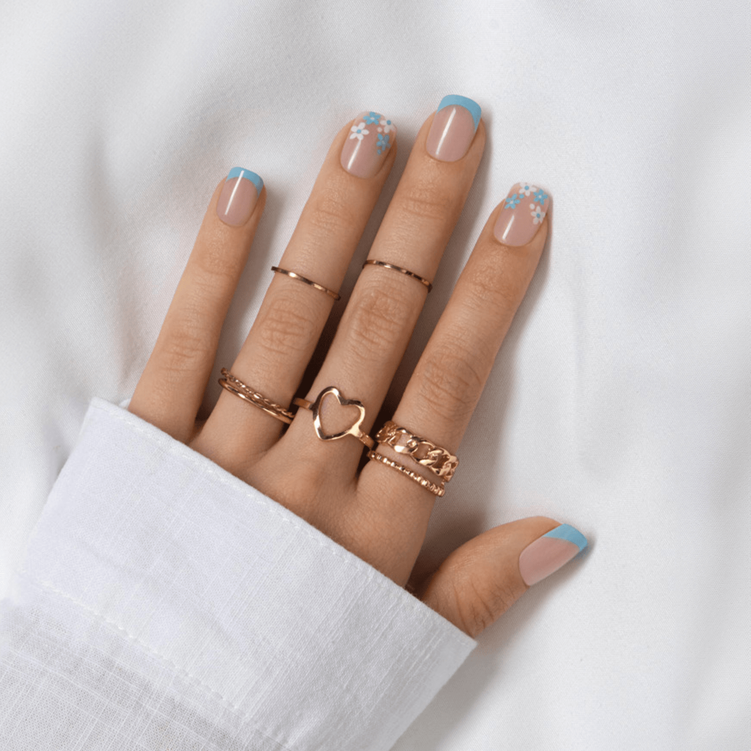 Left hand with five fingers showing French press on nails featuring pale pink base, blue tips, and white and blue floral designs on ring and pinky fingers, adorned with rose gold rings, resting on white fabric.