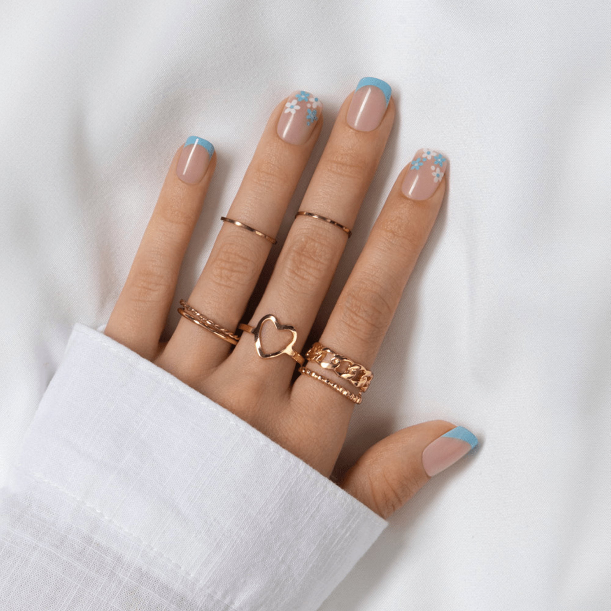 Left hand with five fingers showing French press on nails featuring pale pink base, blue tips, and white and blue floral designs on ring and pinky fingers, adorned with rose gold rings, resting on white fabric.